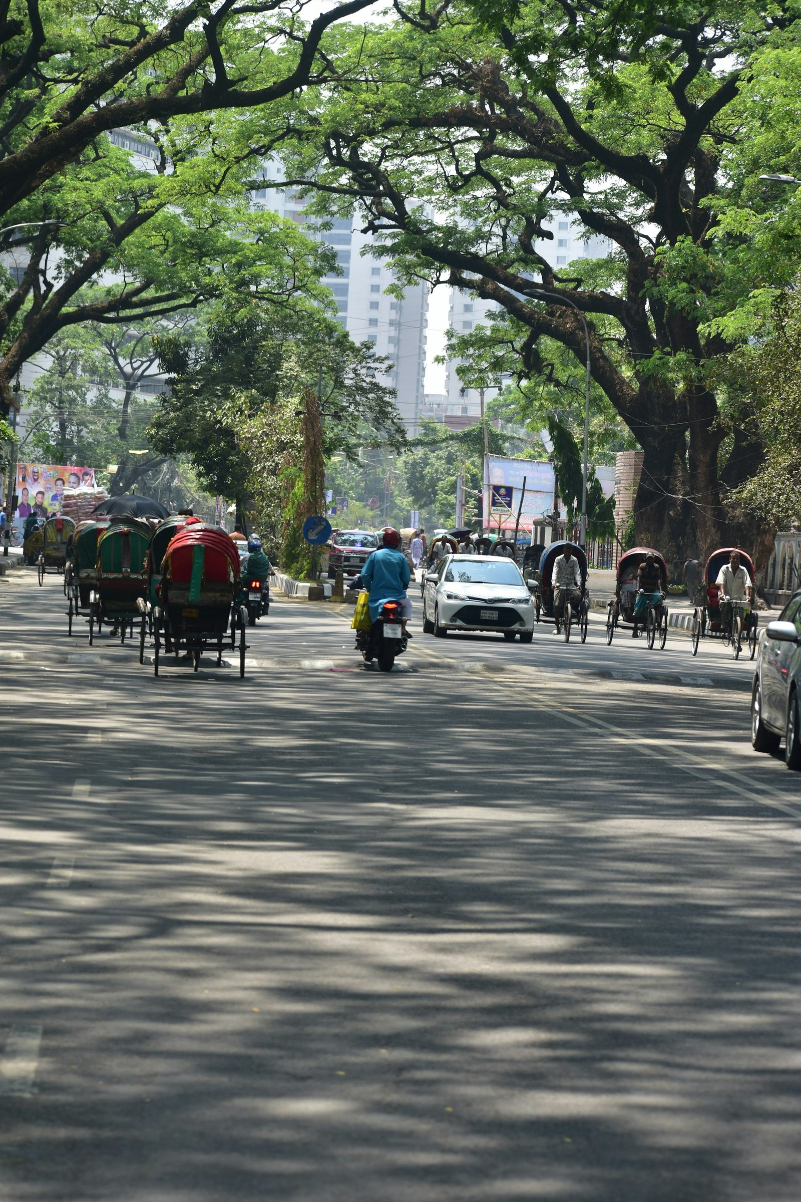 A tree-lined street in Dhaka with rickshaws and traffic